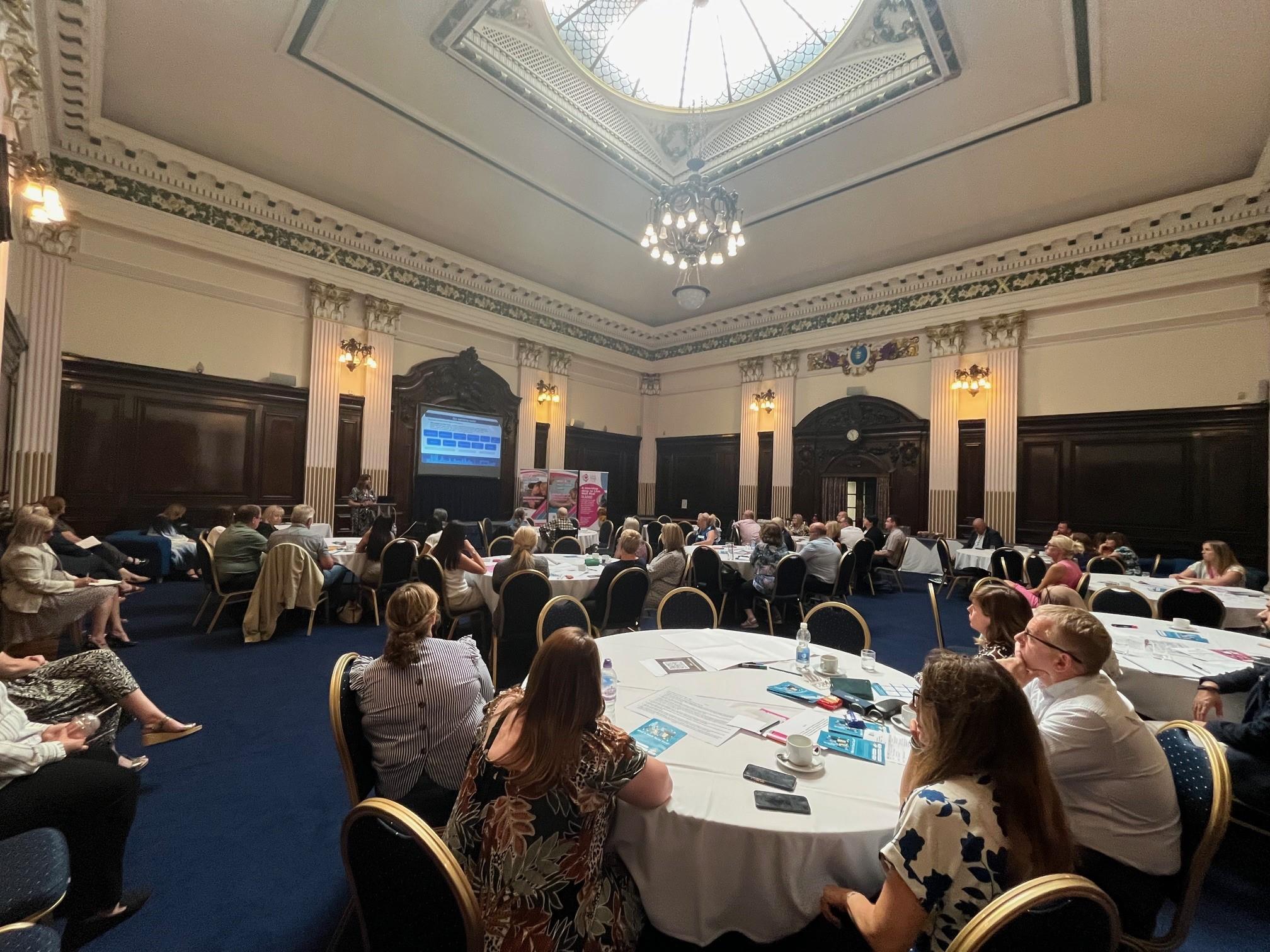 This image shows a picture of Hull Guildhall reception suite. There a people sat around crcukar tables listening to a speaker who is stood at the front of the room giving a presentation.