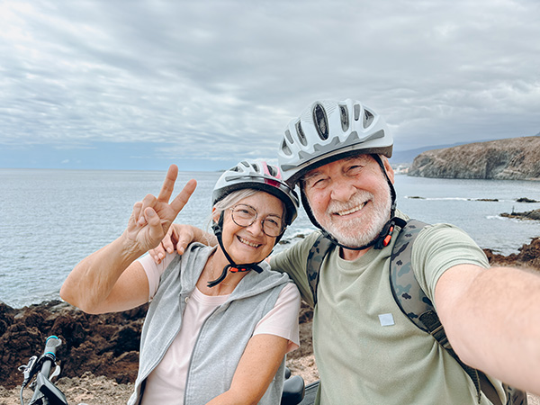 An elderly couple on bikes waving and smiling.